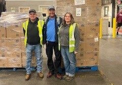 Three individuals standing in a warehouse, positioned in front of stacked boxes, engaged in conversation.