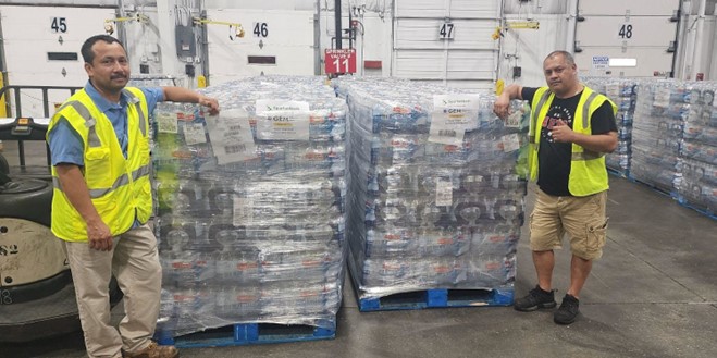 Two men stand beside stacked pallets of bottled water, ready for distribution or storage in a warehouse setting.